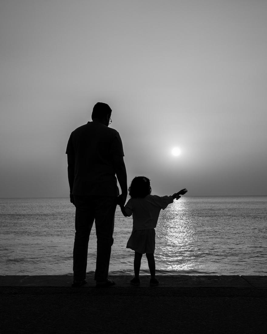 A father and his young child stand hand in hand at the water's edge, watching the sun rise