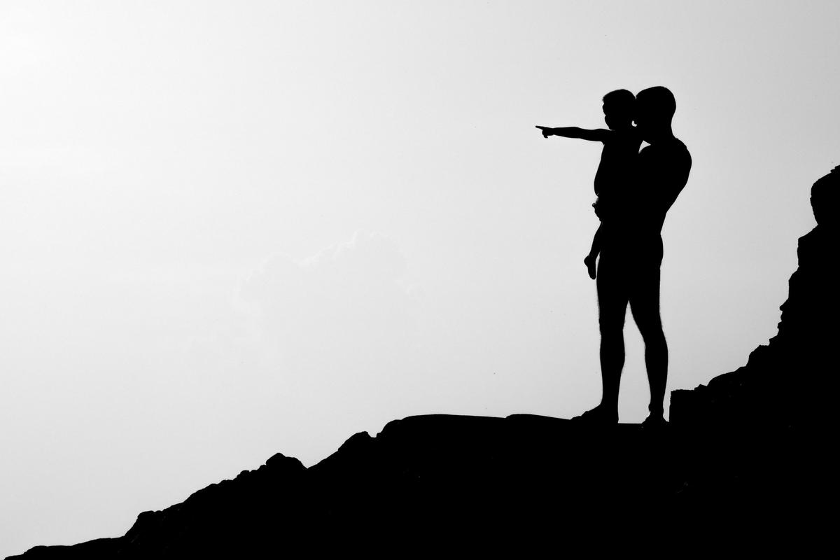 Silhouette of a father on a ridge, holding his child, who points toward the horizon